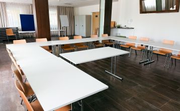 Luchtfoto van Hotel Drei Rosen in Moosburg an der Isar — een vier verdiepingen tellend, lichtpaars hotel met zonnepanelen, glazen balkons en een buitenterras met oranje tafels en stoelen, ideaal voor een voetbaltrainingskamp.