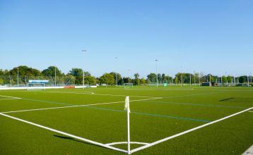 Luchtfoto van Hotel Drei Rosen in Moosburg an der Isar — een vier verdiepingen tellend, lichtpaars hotel met zonnepanelen, glazen balkons en een buitenterras met oranje tafels en stoelen, ideaal voor een voetbaltrainingskamp.