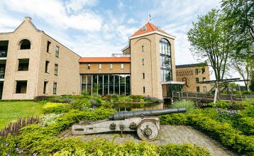 Marveld Recreatie heeft een historisch bakstenen gebouw met een toren, grote ramen en een rood dak in Groenlo. Omgeven door tuinen en bungalows staat een oud kanon op het pad in dit charmante vakantiepark.