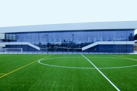 Op de voorgrond ligt het kunstgrasveld Altea, een voetbalveld met wit-gele belijning, terwijl er tegenover een modern gebouw met grote ramen staat.