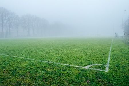 Een mistig voetbalveld met natuurlijke Baarle-Nassau grasmat en witte grenslijnen. Dikke mist verduistert de bomen en doelpalen en creëert een kalme, ingetogen sfeer.