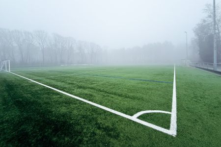 Een mistig voetbalveld met kunstgras Baarle-Nassau, witte grenslijnen en lege doelpalen. Op de mistige achtergrond zijn kale bomen en een hek te zien.