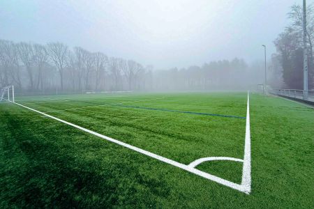Een mistig buitenvoetbalveld met felgroen Baarle-Nassau kunstgras, witte veldmarkeringen en doelpalen aan beide uiteinden, terwijl bladerloze bomen en dichte mist de achtergrond verduisteren.