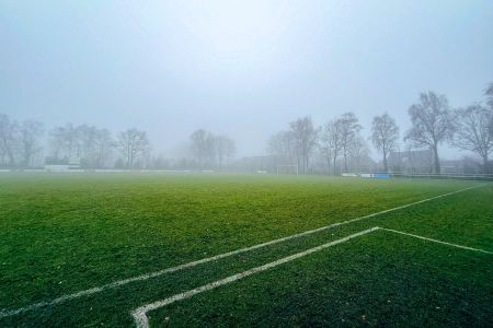 Natuurlijke grasmat 2 Baarle-Nassau: Een mistig voetbalveld met witte grenslijnen, omzoomd met kale bomen en vage contouren van gebouwen op de mistige achtergrond.
