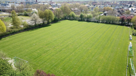 Luchtfoto van trainingsveld 2 Baunatal, een goed onderhouden, leeg voetbalveld met groen gras, duidelijke maailijnen en twee witte doelpalen, omringd door bomen en woongebouwen op een zonnige dag.