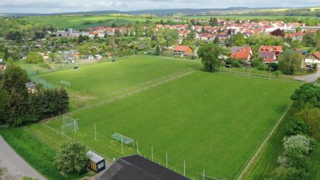 Luchtfoto van natuurgrasveld 2 in Schlotheim, een weelderig groen voetbalveld met twee doelen, omringd door bomen, wegen en een klein stadje met rode daken. Achter de stad liggen tuinen en open velden.