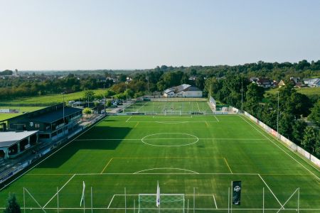 Een luchtfoto van het kunstgrasveld 1 van SC Durbachtal, een leeg groen voetbalveld met witte en gele markeringen, omringd door bomen, gebouwen, geparkeerde auto's, tribunes, hekken en doelpalen aan beide uiteinden onder een heldere hemel.