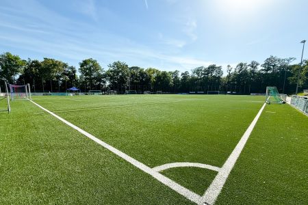 Een heldere, zonnige dag op de Kunstrasenplatz Schöndorf, een leeg kunstgrasvoetbalveld met witte belijning en groene bomen die het veld op de achtergrond omzomen.