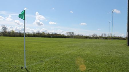Op het natuurgrasveld van Skaerbaek zie je een weelderig groen voetbalveld onder een blauwe hemel met verspreide wolken, een groene hoekvlag op de voorgrond, doelpalen in de verte en hoge lichtmasten aan de rand van het veld.