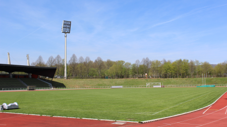 Het stadion van Baunatal heeft een groen veld, een rode atletiekbaan, stadionstoelen, hoge schijnwerpers en bomen op de achtergrond onder een strakblauwe hemel.