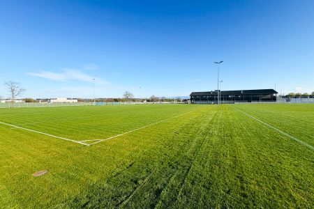 Luchtfoto van trainingsveld Schechen 2, een groen voetbalveld naast een gebouw, omheind en begrensd door velden en een kleine stad, met bergen in de verte onder een strakblauwe hemel.