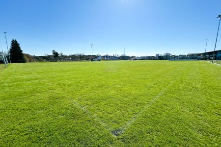 Het trainingsveld van Ebenweiler is een breed, zonovergoten grassportveld met vage witte hoeklijnen. Op de achtergrond zijn bomen, lantaarnpalen en gebouwen te zien onder een strakblauwe hemel.