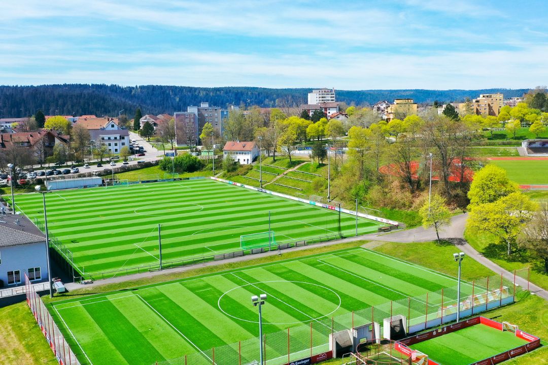 Luftaufnahme der Fußballtrainingsanlage in Baden-Württemberg mit zwei Kunstrasenplätzen, Kleinfeld, Flutlicht und Tribüne, eingebettet in eine grüne Umgebung.