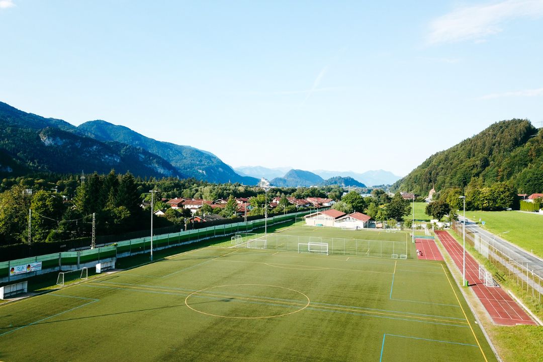 Kunstrasen-Fußballplatz mit einer kurzen Laufbahn daneben in Bayern, eingebettet in eine Alpenlandschaft mit malerischem Dorf im Hintergrund.