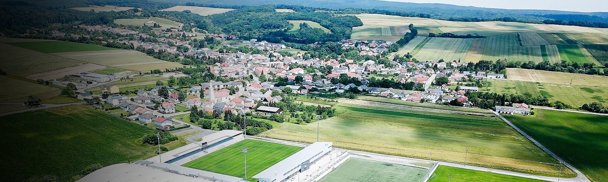 Luchtfoto van een sportcentrum in Burgenland, omringd door groene velden en een klein dorp met rode daken.