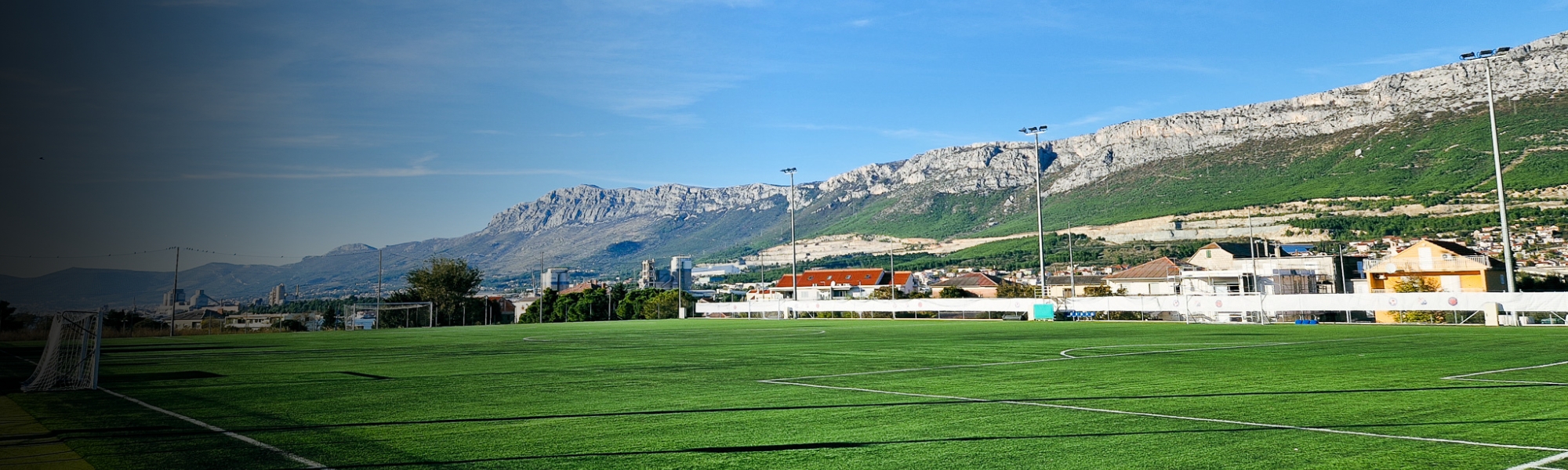 Voetbalveld in Dalmatië met schijnwerpers en een indrukwekkend bergpanorama op de achtergrond, onder een strakblauwe hemel.