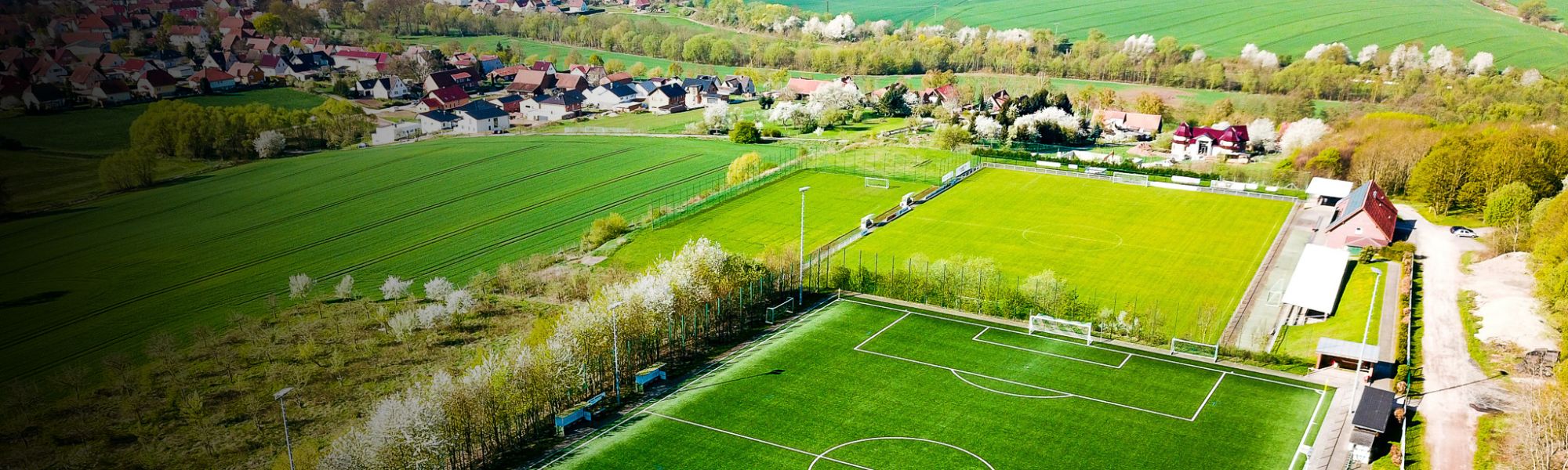 Panoramische luchtfoto van een voetbalfaciliteit met een kunstgrasveld en een natuurgrasveld, ingebed in een landelijk landschap met bloeiende bomen, naburige velden en een klein dorp op de achtergrond.