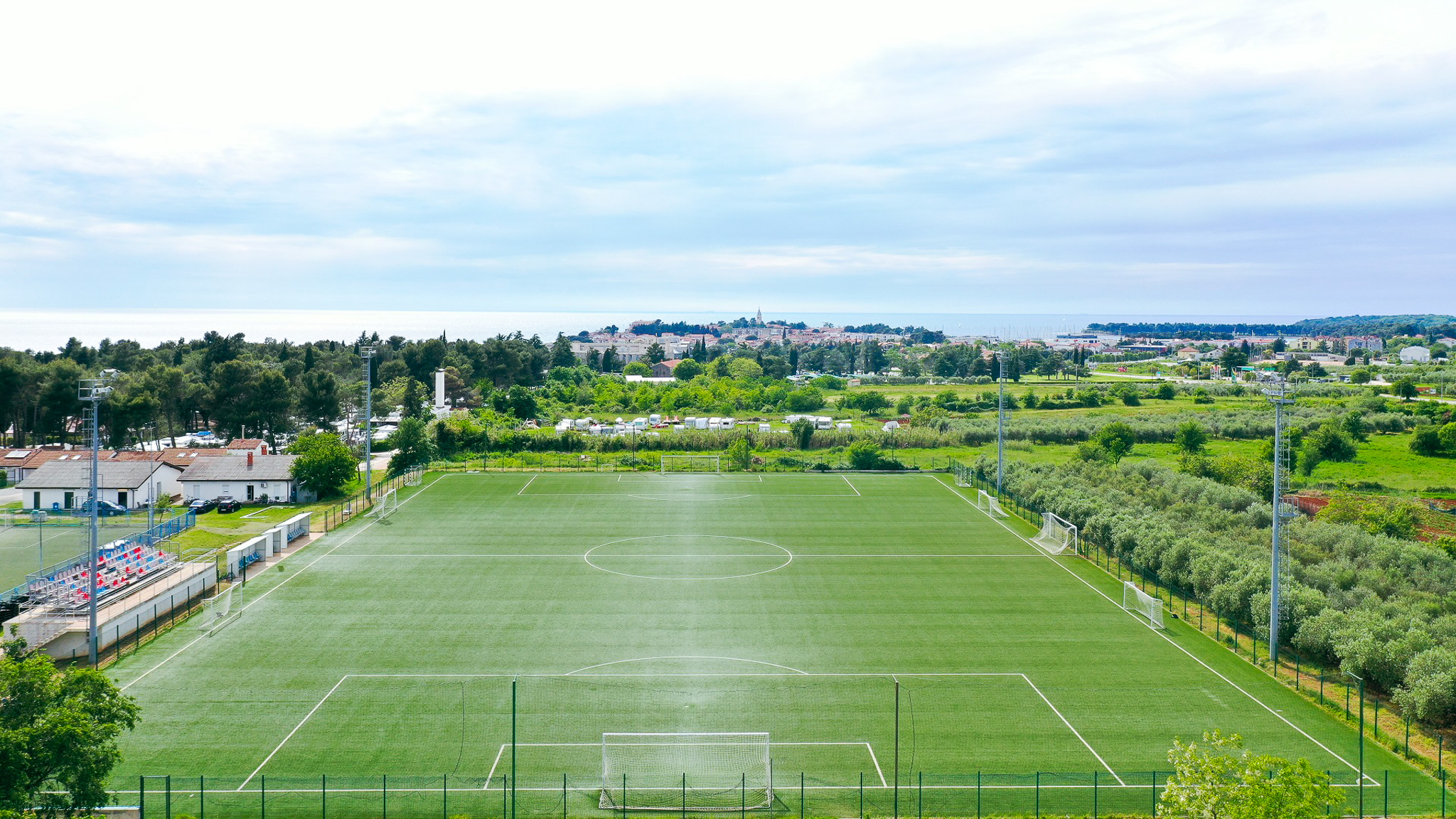 Voetbalveld in Istrië met kunstgras, omgeven door groene natuur en olijfbomen, met de zee en de stad Poreč op de achtergrond.