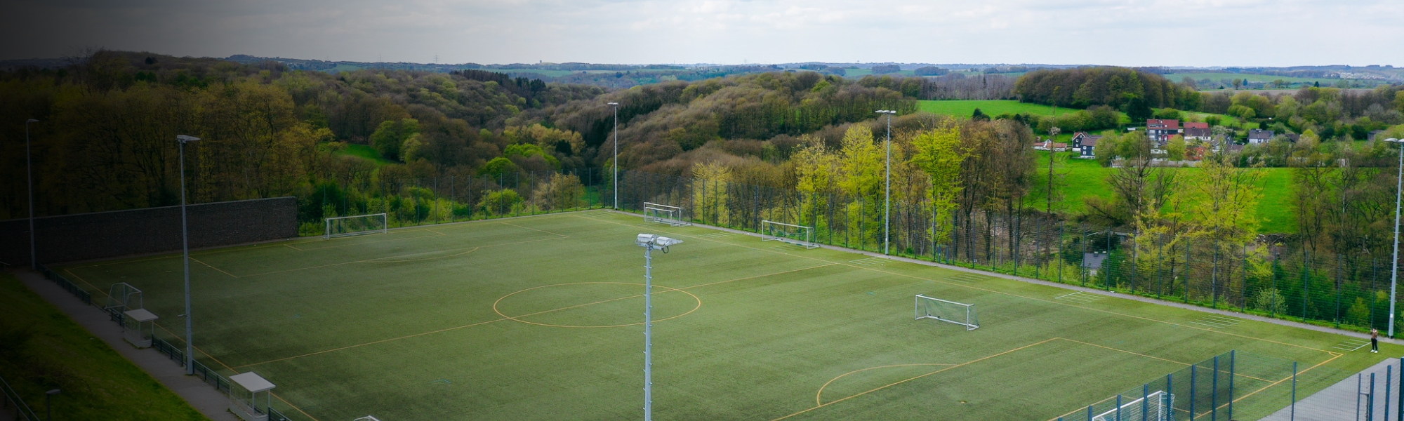 Uitgestrekt voetbaltrainingsveld met kunstgrasveld in Noordrijn-Westfalen, ingebed in een schilderachtig natuurlandschap met dichte bossen en heuvels.