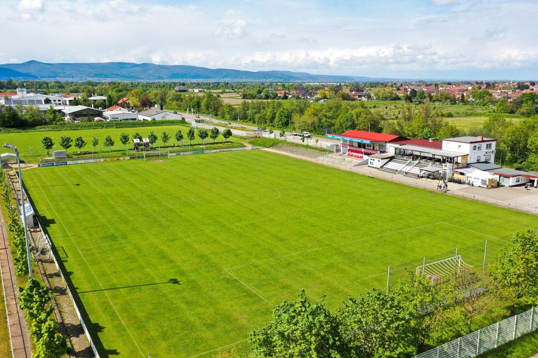 Luchtfoto van een goed onderhouden voetbalveld van natuurgras met aangrenzend clubhuis en tribune, ingebed in een groen landschap met uitzicht op een stad en heuvels op de achtergrond in Rijnland-Palts.