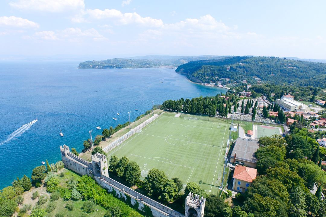 Fußballtrainingsplatz direkt am Meer in Slowenien, mit Blick auf die Küste, die umliegenden Hügel und ein kleines Dorf mit typischen südeuropäischen Gebäuden.
