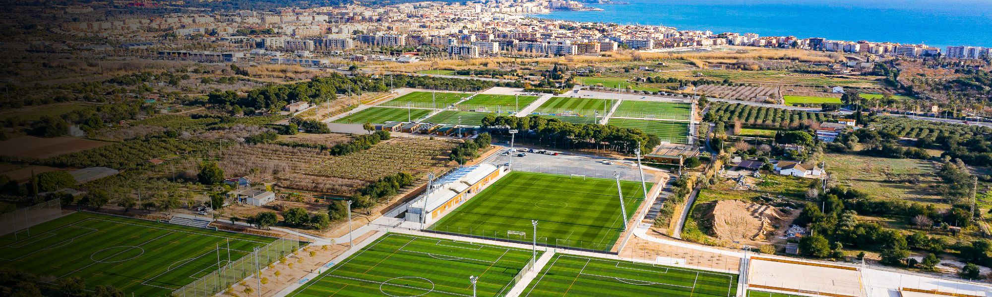 Panoramische opname van een grote voetbaltrainingsfaciliteit in Spanje, bestaande uit verschillende natuurgras- en kunstgrasvelden, omringd door landbouwvelden, olijfgaarden en een kustplaats met de Middellandse Zee op de achtergrond.