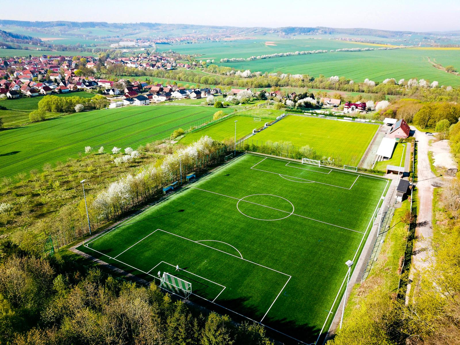 Luftaufnahme einer modernen Fu&szlig;ballanlage mit einem Kunstrasen- und einem Naturrasenplatz, umgeben von bl&uuml;henden B&auml;umen, Feldern und einem kleinen Dorf in einer l&auml;ndlichen H&uuml;gellandschaft.