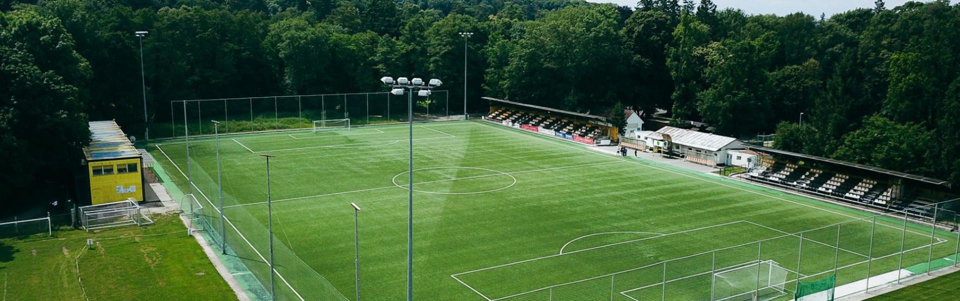 Fotballtreningsbane omgitt av tette skoger, med en liten tribune og flomlysmaster.
