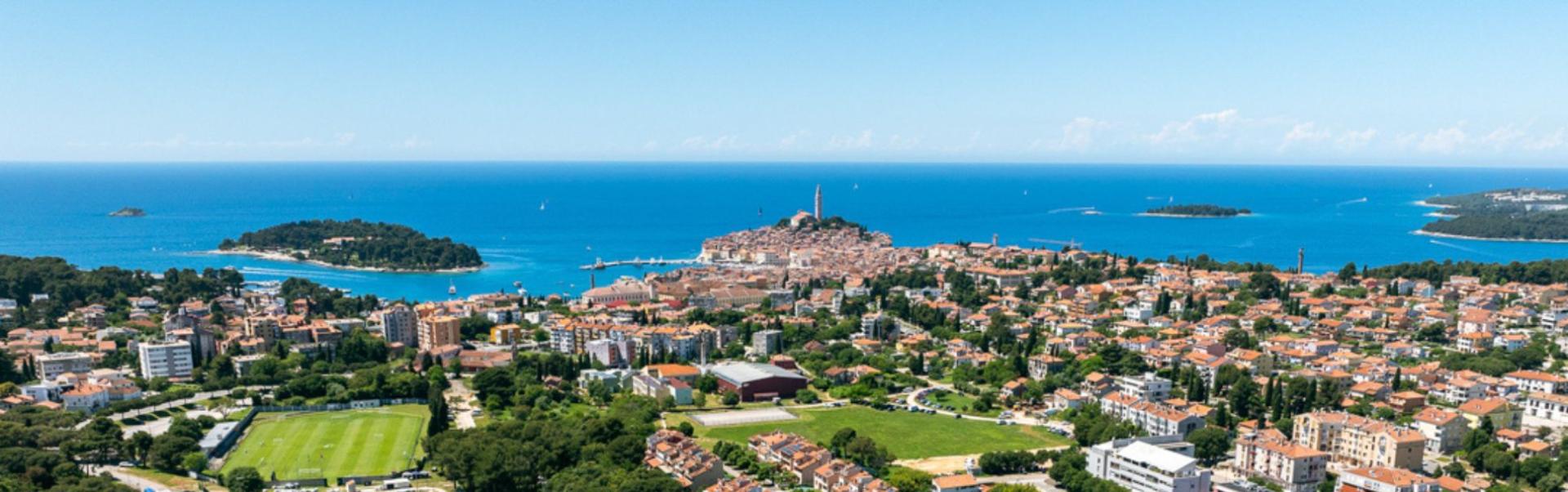 Panoramisk luftfoto av et fotballtreningsanlegg i Kroatia med flere baner med naturgress og kunstgress, flomlysmaster, omgitt av bolighus og gr&oslash;ntomr&aring;der, med en liten by og Adriaterhavet i bakgrunnen.