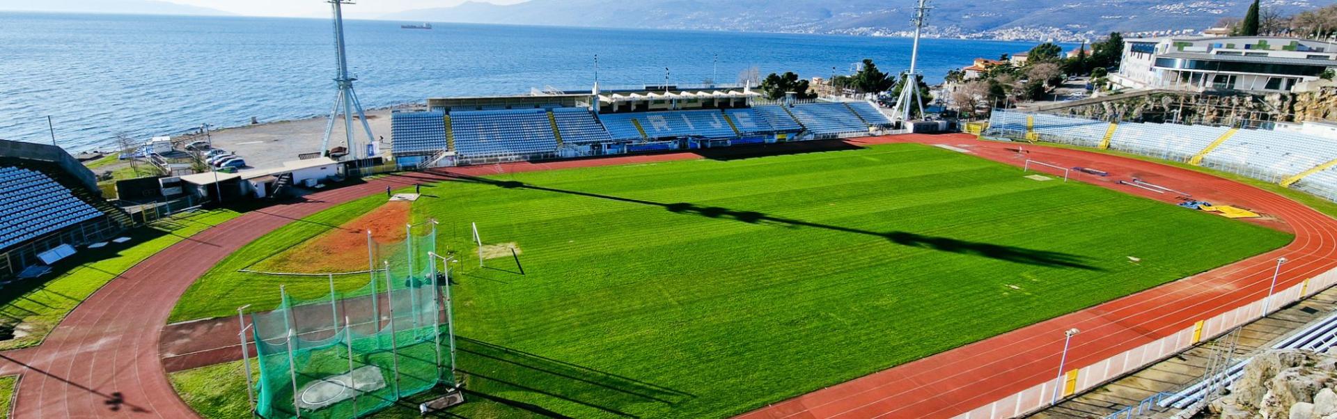 Luftfoto av en fotballstadion ved Kvarnerbukten i Kroatia, beliggende rett ved sj&oslash;en, med l&oslash;pebane og tribuner, omgitt av fjell og Adriaterhavet i bakgrunnen.