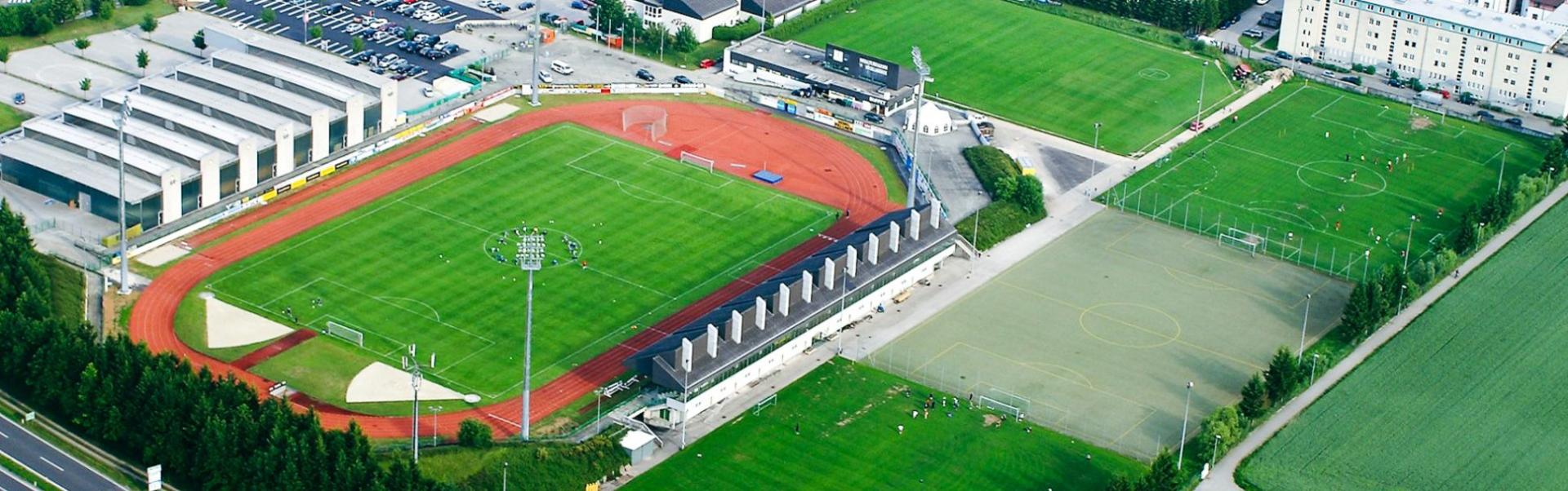 Luftfoto av et moderne idrettsanlegg i Ober&ouml;sterreich med flere fotballbaner, blant annet et stadion med l&oslash;pebane, kunstgressbaner og naturgressbaner, omgitt av parkeringsplasser, n&aelig;ringsbygg og bolighus.