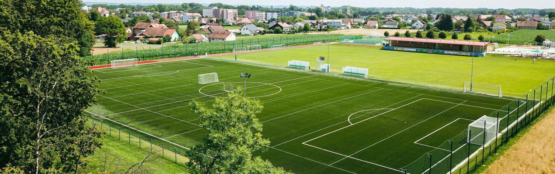 Fotballtreningsleir med kunstgressbane og naturgressbane midt i et idyllisk landskap med sm&aring; hus i bakgrunnen.