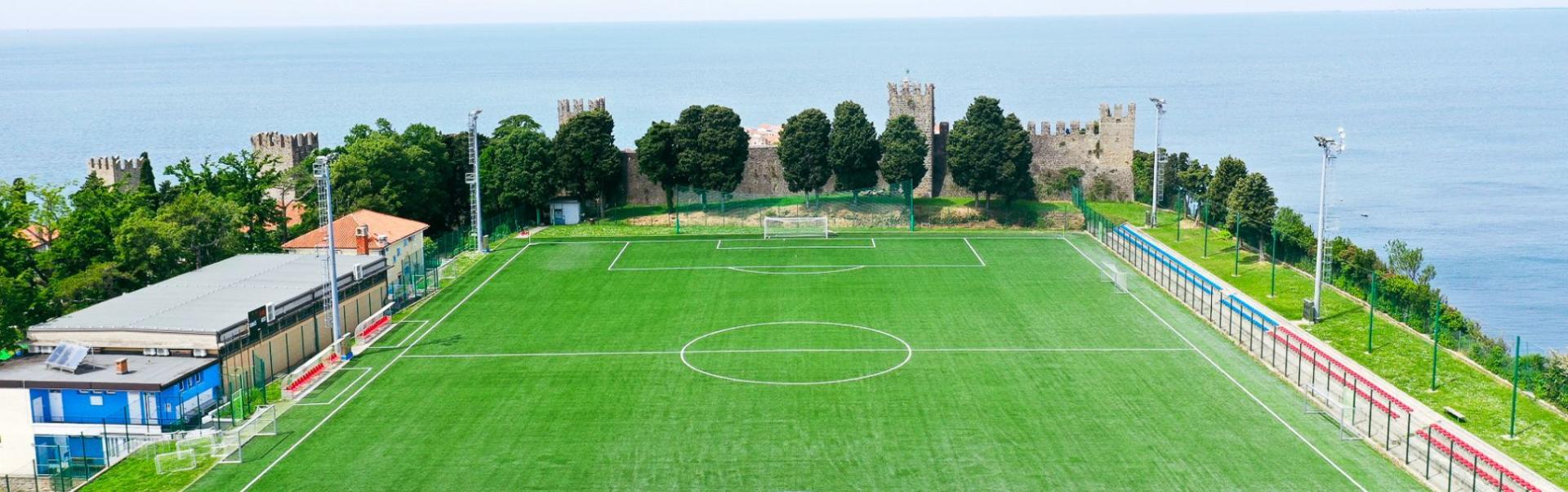  Luftfoto av en fotballbane ved kysten, omgitt av gr&oslash;nt landskap og bl&aring;tt hav.