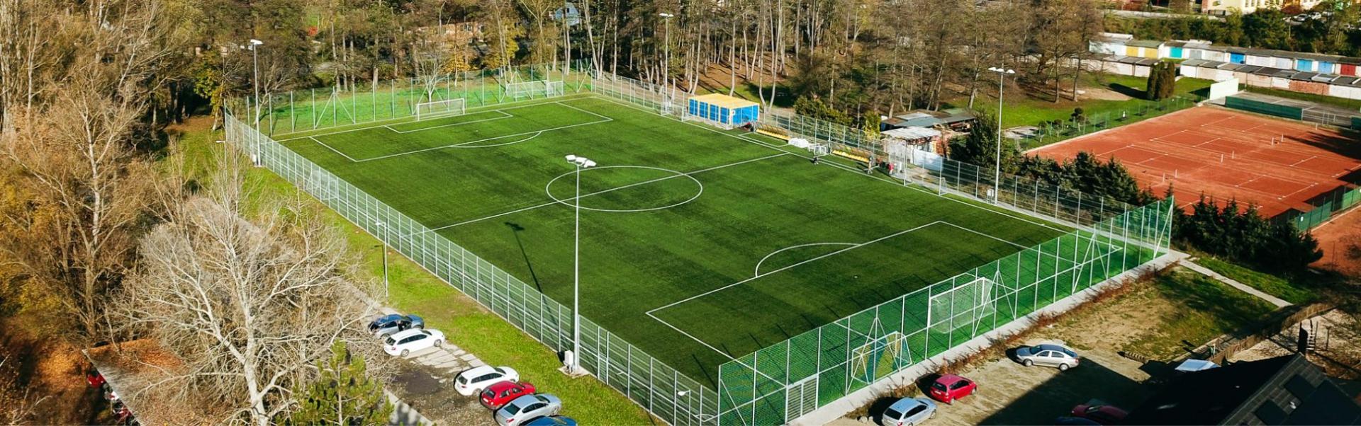Fotballbane med kunstgress, beliggende i en liten by, omgitt av skog og tennisbaner.