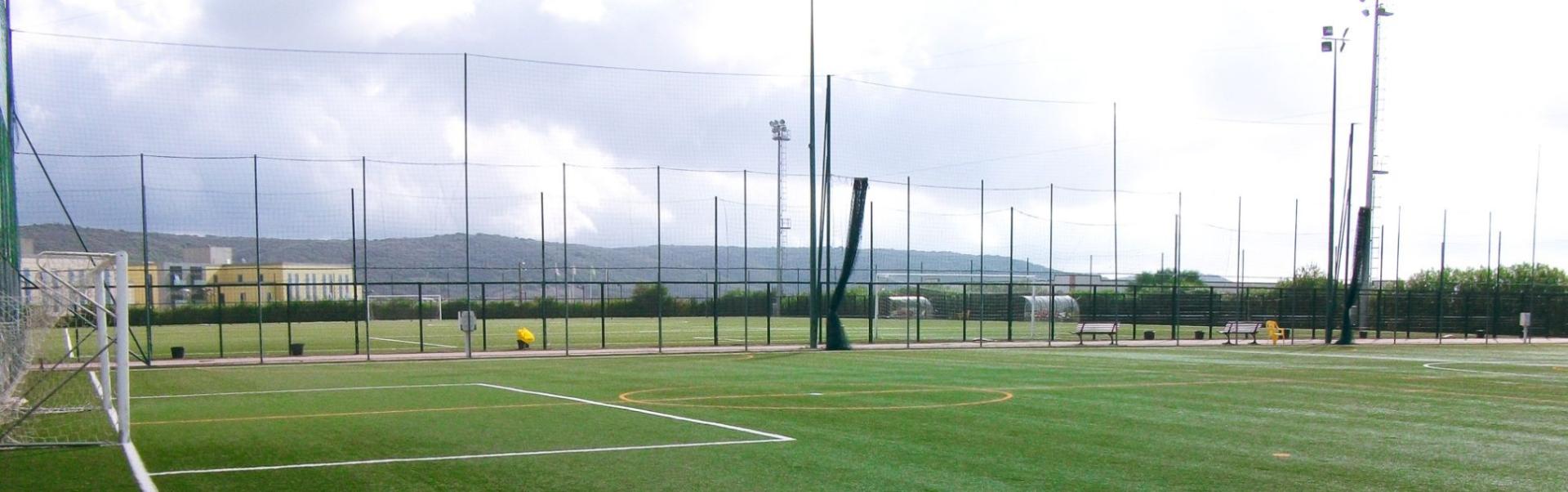 Fotballtreningsbane p&aring; Sardinia med gr&oslash;nt kunstgress, m&aring;l, benker og flomlysmaster, omgitt av nett og fjellandskap.