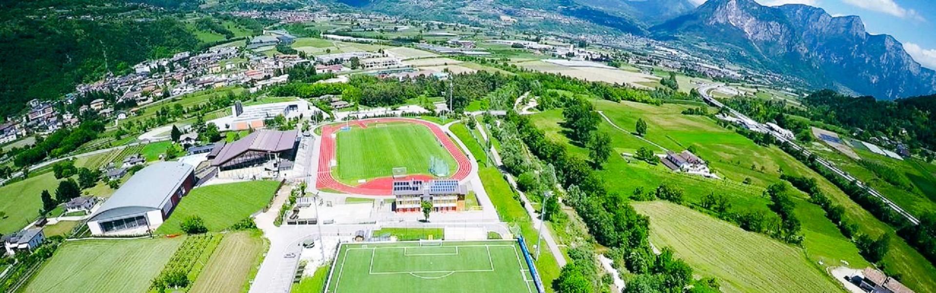 Luftfoto av en moderne fotballbane med tribune og tilst&oslash;tende friidrettsanlegg midt i det gr&oslash;nne landskapet i Trentino, omgitt av fjell og sm&aring; landsbyer.