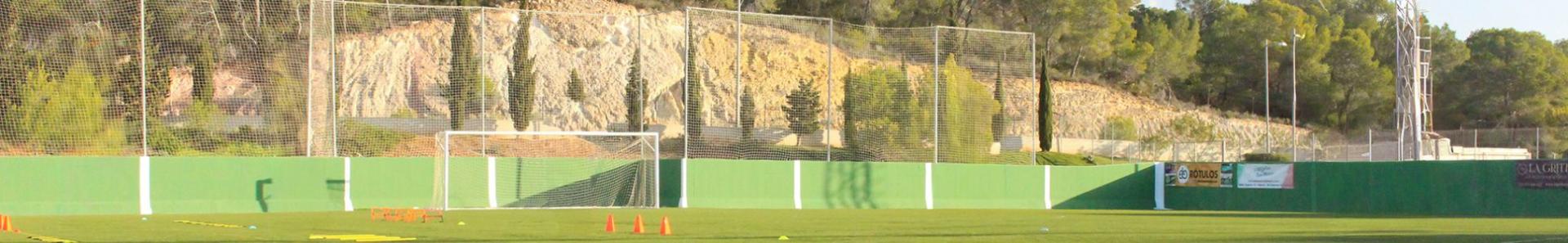 En fotballbane med gr&oslash;nt gjerde, oransje treningskegler, stiger, nett bak m&aring;let og tr&aelig;r og steinete &aring;ser i bakgrunnen under en klar himmel.