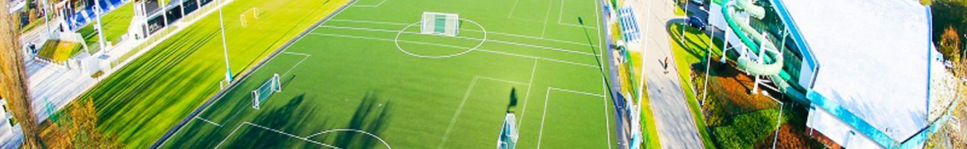 Luftfoto av et moderne fotballtreningsanlegg med flere baner, tribuner og en tilst&oslash;tende idrettshall