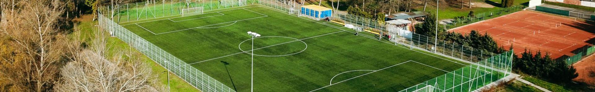 Fotballbane med kunstgress, beliggende i en liten by, omgitt av skog og tennisbaner.