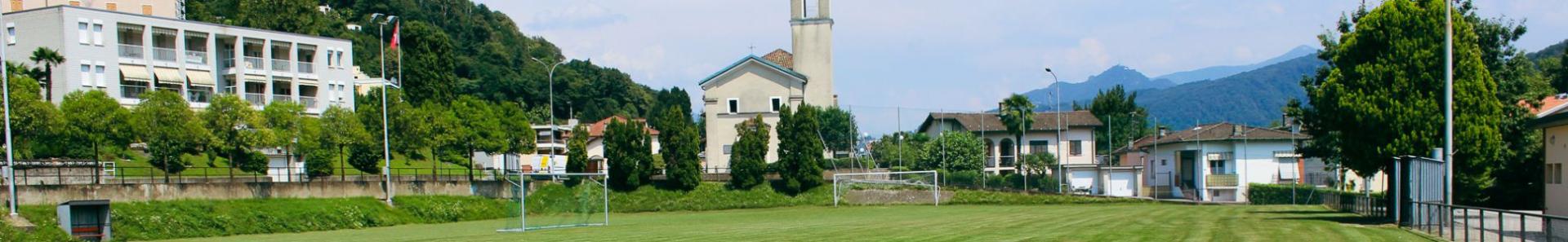 En velstelt fotballbane i den pittoreske regionen Ticino, omgitt av grønne åser og tradisjonell arkitektur. I bakgrunnen ligger en kirke og bolighus som danner en idyllisk kulisse for et fotballtreningsleir.