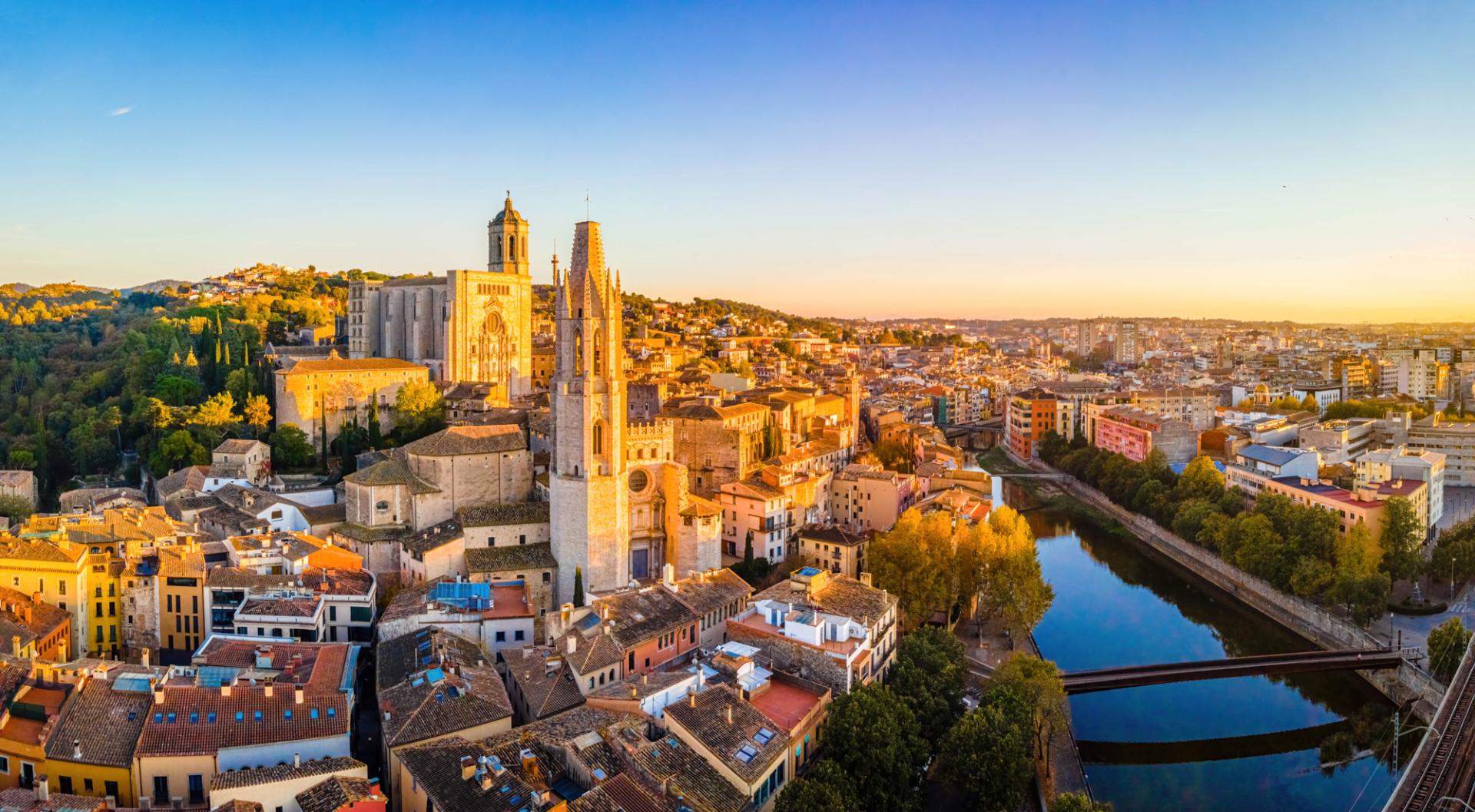 Die Altstadt von Girona in Spanien bei Sonnenuntergang, mit beeindruckenden gotischen und romanischen Bauwerken, umgeben von modernen Stadtteilen und einem ruhigen Fluss, der durch die Stadt flie&szlig;t