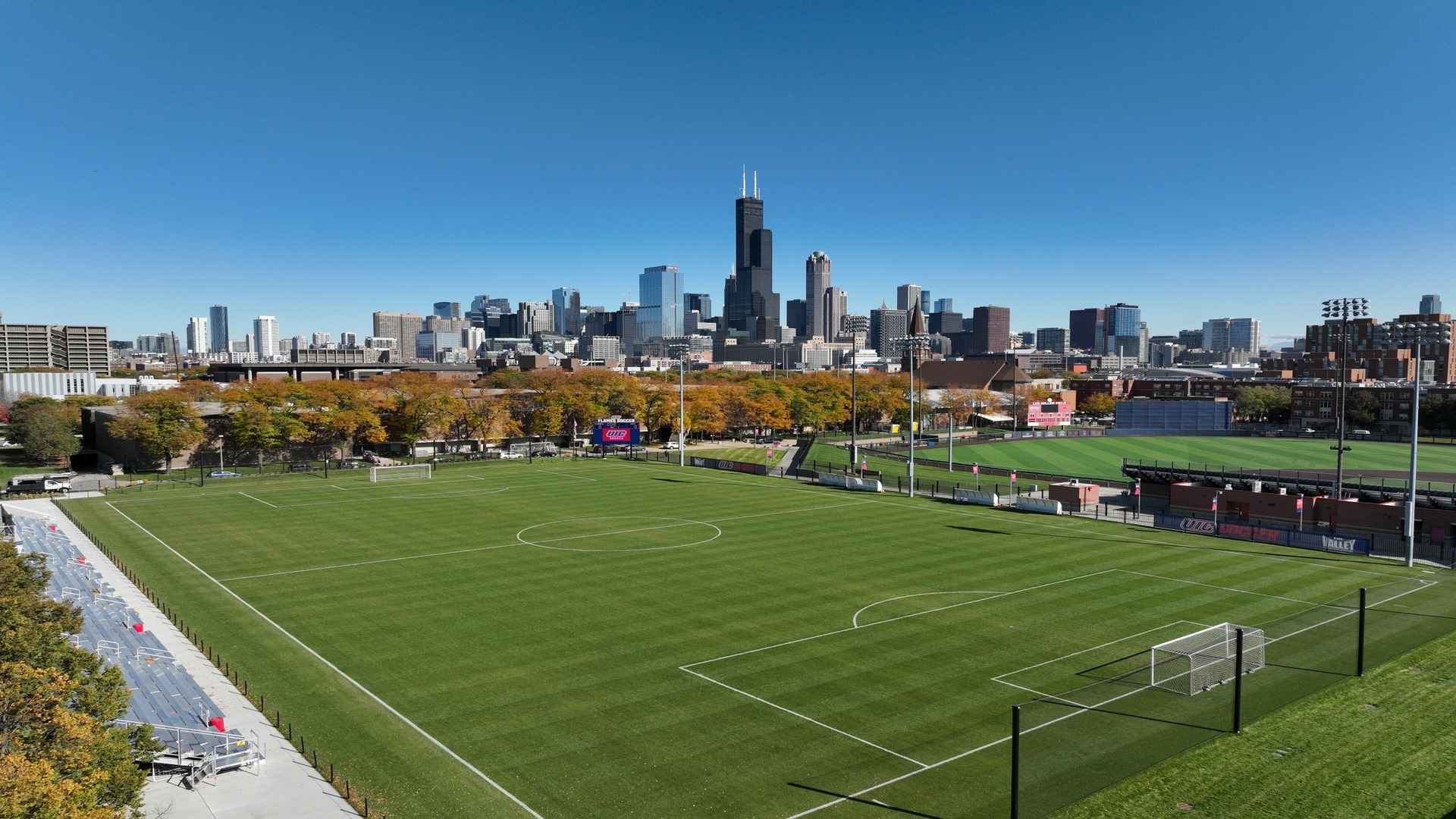 Fu&szlig;ballfeld mit gepflegtem Rasen vor einer modernen Skyline, umgeben von Flutlichtmasten und Trib&uuml;nen unter klarem, blauem Himmel.
