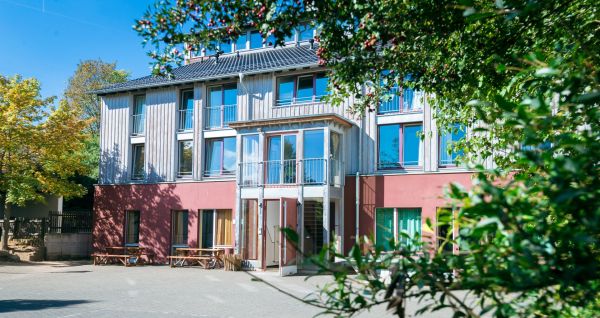 Das Gästehaus Nettersheim ist ein modernes, dreistöckiges Gebäude mit großen Fenstern, einer roten unteren Fassade, hölzernen Obergeschossen und einem kleinen Balkon, eingebettet in eine grüne, natürliche Umgebung mit Bäumen unter der Sonne.