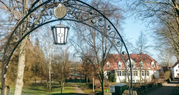 Ein verschnörkelter schwarzer Metallbogen mit einer hängenden Laterne steht am Eingang des Parks in der Nähe eines weißen Gebäudes mit rotem Ziegeldach und Bäumen - ideal für Besucher, die zum Gästehaus Northeim wollen oder auf der Suche nach nahe gelegen
