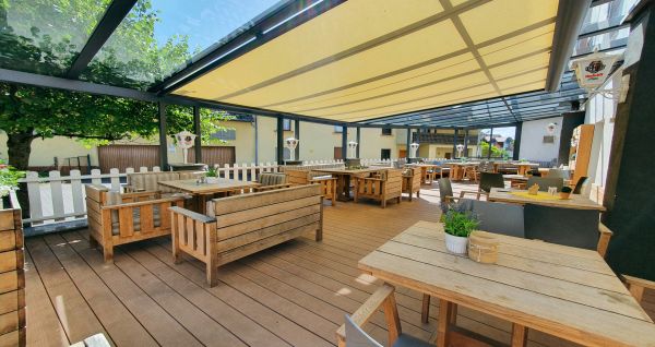 Das Landhotel Imhof verfügt über eine geräumige Außenterrasse mit Holztischen und -stühlen unter einem gelben Baldachin, Topfpflanzen auf den Tischen und einem weißen Lattenzaun, der für eine helle und entspannte Atmosphäre beim Essen sorgt.