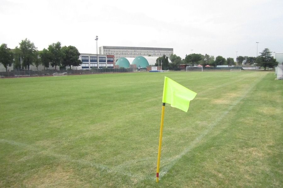 Eine leuchtend gelbe Eckfahne steht auf einem Rasenfußballfeld mit entfernten Torpfosten; im Hintergrund sind Bäume, Gebäude und die Appartements Residence Eden*** unter einem bewölkten Himmel zu sehen.
