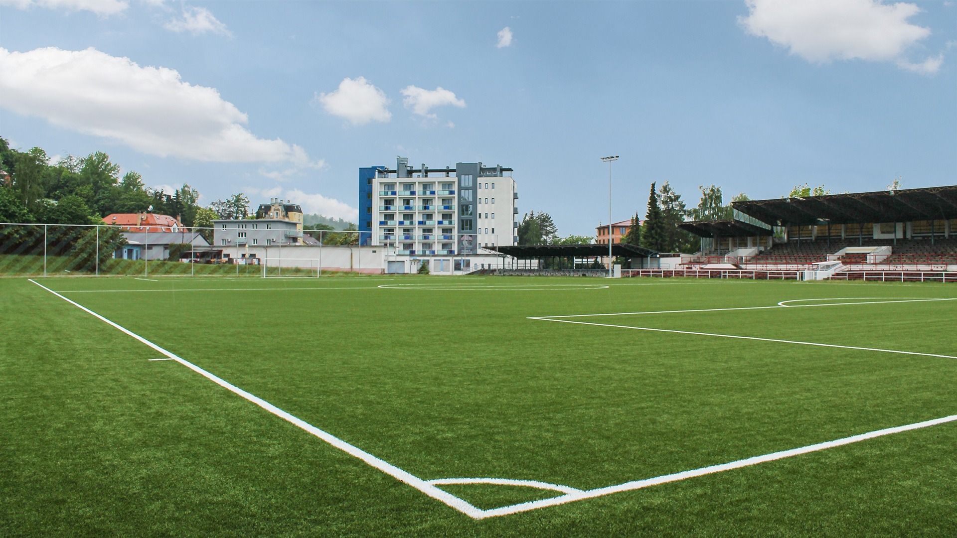 Ein grüner Kunstrasen-Fußballplatz mit weißen Linien liegt neben Tribünen und dem mehrstöckigen Hotel Marttel*** - einem blau-weißen Gebäude - vor einem blauen Himmel mit vereinzelten Wolken.