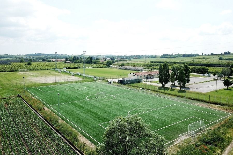 Luftaufnahme eines grünen Fußballplatzes mit Toren, ideal für preisbewusste Mannschaften, umgeben von Bäumen, Ackerland und Appartements Residence Eden*** in der Nähe. Sportplätze und Parkplätze sind unter einem teilweise bewölkten Himmel sichtbar.