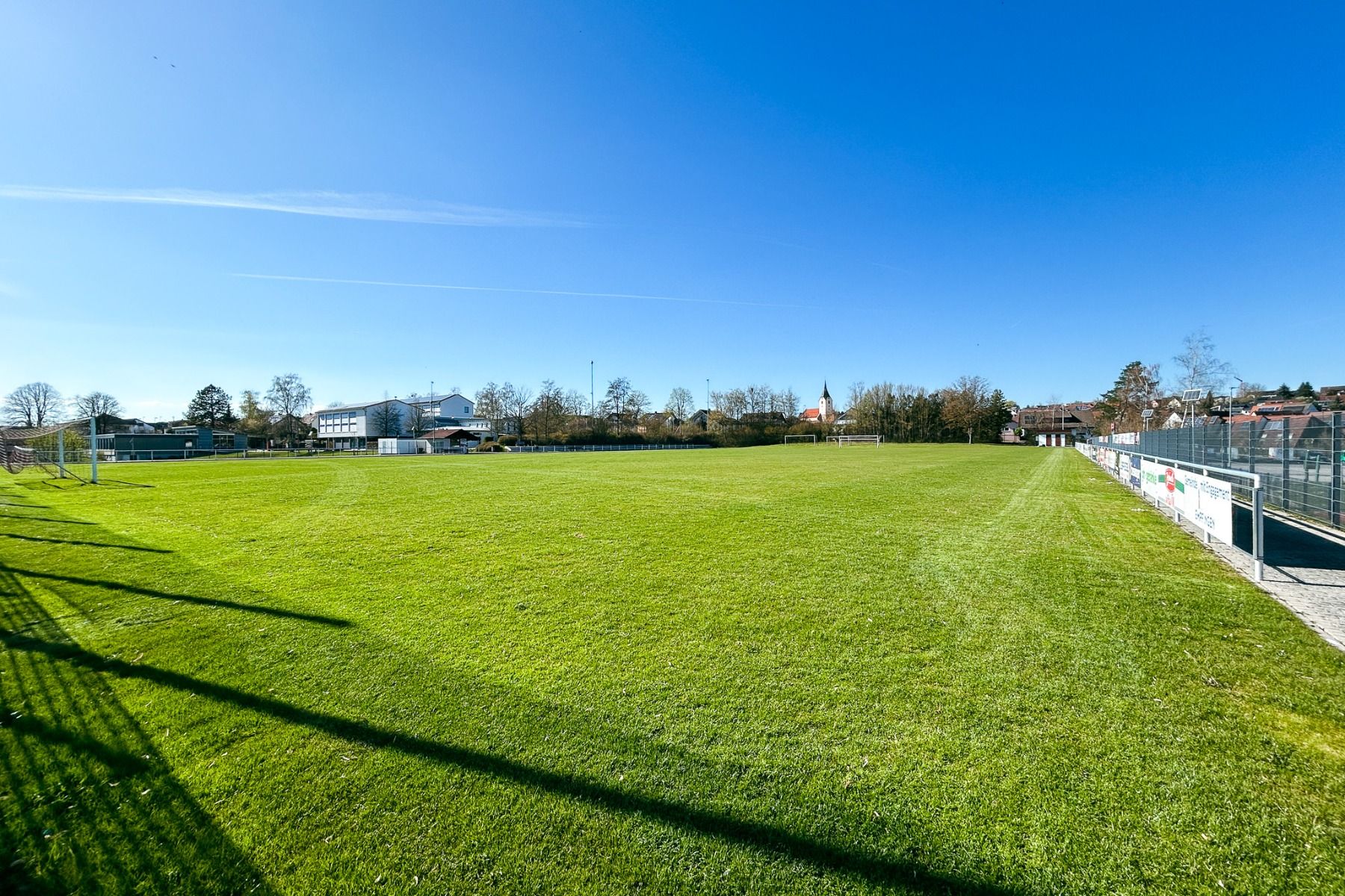 En bred, grønn fotballbane under en knallblå himmel - ideell for en fotballtreningsleir - er inngjerdet og omkranset av bygninger og trær i nærheten av det komfortable Hotel Empfinger Hof på en solrik dag.