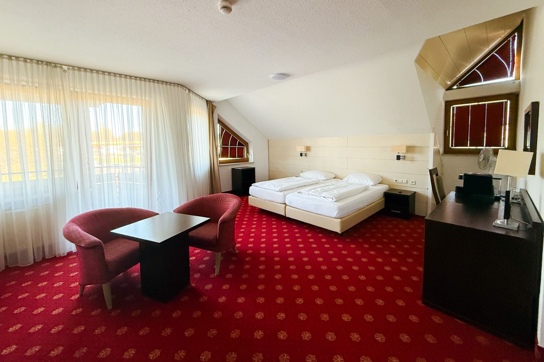 Et koselig rom på Hotel Empfinger Hof har to enkeltsenger, rødt teppe, to røde lenestoler ved et lite bord, et skrivebord med speil, store vinduer med skjørtgardiner og skråtak - ideelt for et opphold i et fotballtreningslager.
